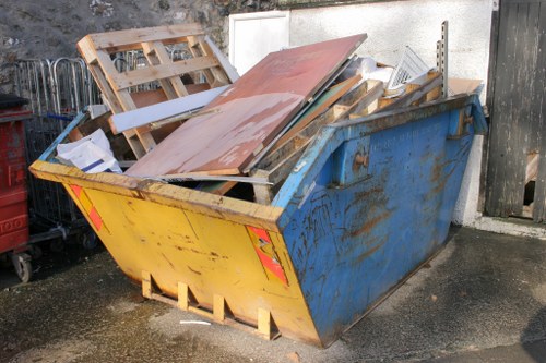 A large, blue metal skip positioned on an asphalt surface outdoors, filled with various discarded items including wooden pallets, sheets of metal and wood, and other miscellaneous waste materials. The skip shows signs of rust and weathering, with the top layer of materials haphazardly piled, some leaning over the edges. The background features a concrete wall and a weathered wooden fence, with the scene suggesting a typical rubbish removal operation in a residential or commercial area within the vicinity of Seven Sisters, London. The image captures the practicality of waste clearance by Seven Sisters House Clearance, highlighting the type of bulky refuse managed by professional rubbish removal services. The environment is well-lit with natural light, emphasizing the textures and colours of the debris and metal container.
