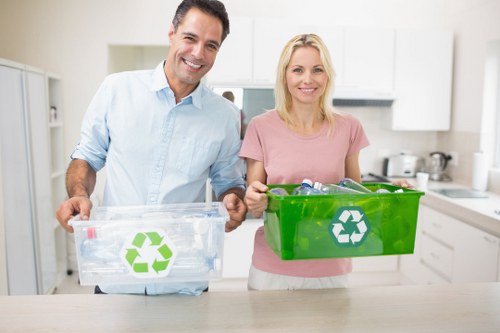 Two smiling individuals, a man in a light blue shirt and a woman in a pink top, are standing in a modern, bright kitchen. They are holding transparent and green recycling boxes with white recycling symbols on the front, suggesting they are about to sort waste. The kitchen features white cabinets, a white backsplash, and a stainless steel kettle on the countertop in the background. The setting appears clean and organized, with natural lighting illuminating the scene. The image is relevant to rubbish and waste management services offered by companies such as Seven Sisters House Clearance, emphasizing environmentally conscious waste separation and recycling practices, possibly within the London postcode area. The focus is on responsible waste disposal and recycling, aligning with professional rubbish removal services locally provided in the area.