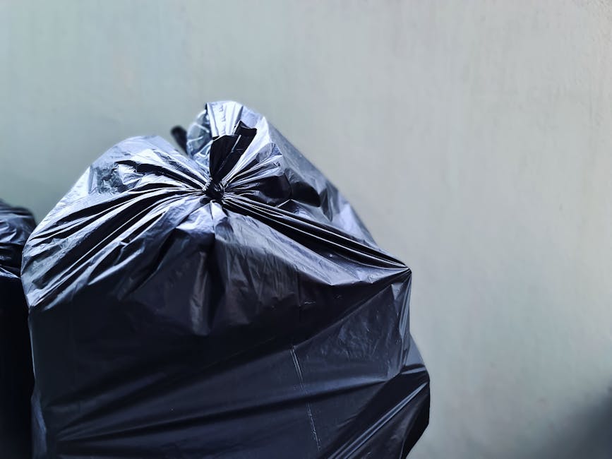 Close-up view of a black plastic rubbish bag with a tightly knotted top, sitting on or next to other similar black bags. The bags are made of shiny, crinkled plastic material and appear to be filled w