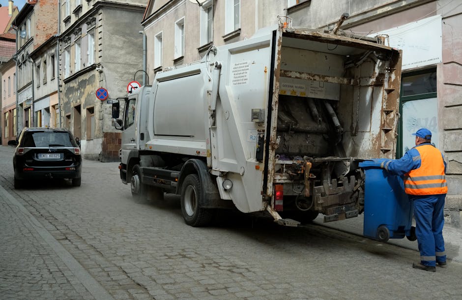 A narrow urban street scene showing several workers in high-visibility clothing and helmets gathered around a black wheelie bin and a clutter of discarded waste items on the pavement. The alleyway is 